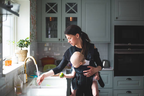 Mother washing hand in kitchen sink while carrying baby girl at home - MASF13436