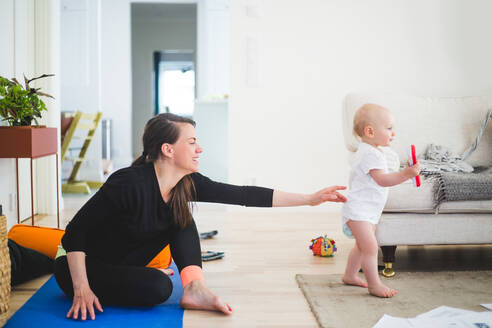 Seitenansicht einer berufstätigen Mutter, die nach ihrer Tochter greift, während sie auf einer Gymnastikmatte im Wohnzimmer zu Hause sitzt - MASF13442