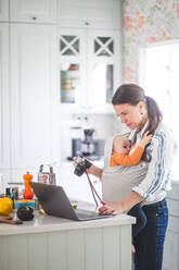 Seitenansicht eines Food-Bloggers mit Kamera und Laptop, während er ein kleines Mädchen in der Küche trägt - MASF13464