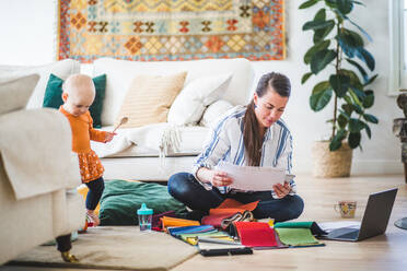 Modedesignerin prüft Papiere, während ihre Tochter im Wohnzimmer spielt - MASF13472