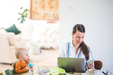 Baby-Mädchen schaut auf beschäftigte arbeitende Mutter, während sie am Tisch im Heimbüro sitzt, lizenzfreies Stockfoto