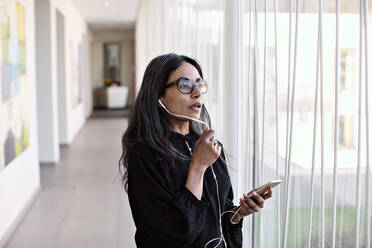 Geschäftsfrau, die mit ihrem Smartphone telefoniert, während sie am Fenster im Büroflur steht - MASF13492
