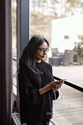 Businesswoman using smart phone while standing by window in office - MASF13503