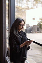 Geschäftsfrau, die ein Smartphone benutzt, während sie am Fenster im Büro steht - MASF13503