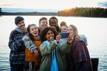 Happy male and female friends taking selfie with smart phone while standing against lake during sunset - MASF13561