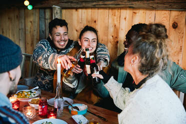 Happy male and friends toasting beer bottles while sitting in log cabin - MASF13577