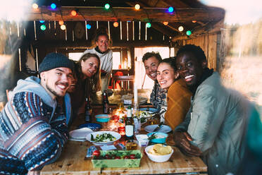 Porträt von lächelnden Freunden mit Essen und Getränken auf dem Tisch in einem Landhaus, gesehen durch ein Fenster - MASF13582