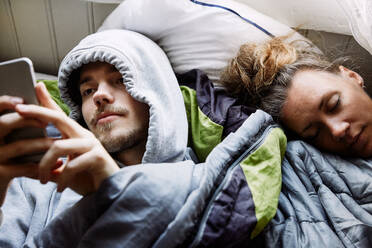 Hoher Blickwinkel eines jungen Mannes, der sein Smartphone benutzt, während er zu Hause neben einem Freund auf dem Bett liegt - MASF13605