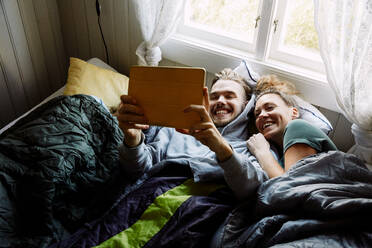 High angle view of smiling friends watching movie over digital tablet while lying on bed in cottage - MASF13611