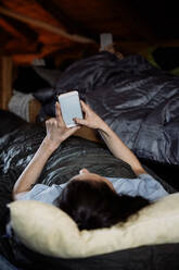 Junge Frau, die ihr Smartphone benutzt, während sie auf einer Matratze in einer Hütte liegt - MASF13616