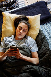 High angle view of young woman using smart phone while lying on mattress in cottage - MASF13617