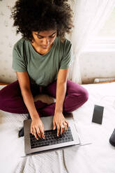 High angle view of young woman with curly hair using laptop while sitting on bed at home - MASF13627