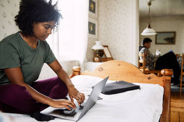 Junge Frau, die einen Laptop auf dem Bett benutzt, während ein Freund im Hintergrund zu Hause sitzt - MASF13628