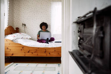 Junge Frau, die einen Laptop benutzt, während sie sich zu Hause auf dem Bett entspannt, gesehen durch die Türöffnung - MASF13630