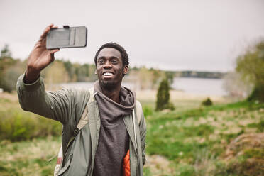 Lächelnder junger Mann, der ein Selfie mit seinem Smartphone macht, während er auf einer Landschaft gegen den Himmel steht - MASF13632