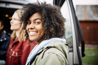 Portrait of smiling young woman with curly hair standing with friend by car - MASF13648