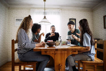 Happy friends talking while enjoying food and drink on table at home - MASF13658