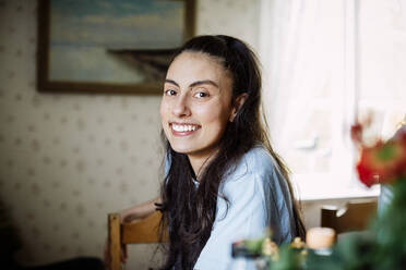 Portrait of smiling young woman with long hair sitting on chair at home - MASF13659