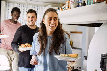 Lächelnde Freunde, die Essen in der Hand halten, während sie in der Küche zu Hause stehen - MASF13663