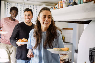 Smiling friends holding food while standing in kitchen at home - MASF13663