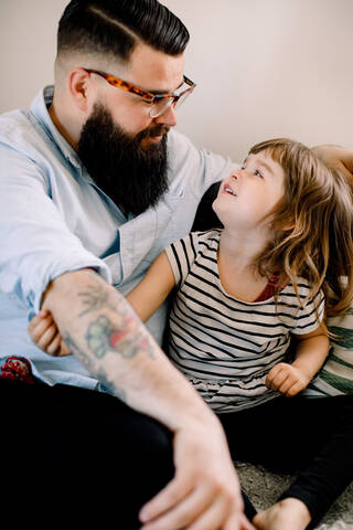 Lächelnder Vater im Gespräch mit seiner Tochter, während er zu Hause auf dem Sofa sitzt, lizenzfreies Stockfoto