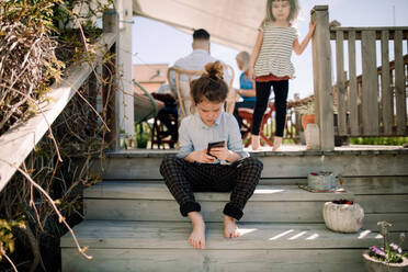 Girl using mobile phone while sitting on steps with sibling in background at patio - MASF13690