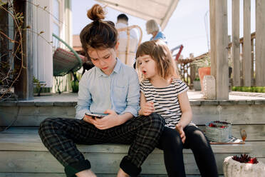 Girl talking to sister using smart phone while sitting on steps in patio - MASF13692