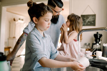 Mädchen wäscht Tasse am Waschbecken, während der Vater mit der Tochter in der Küche spricht - MASF13703