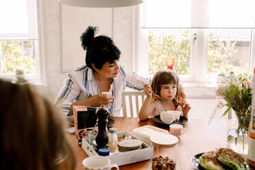 Mother looking at daughter having breakfast on dining table - MASF13712