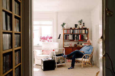 Full length of senior man sitting on chair in living room seen from doorway - MASF13716