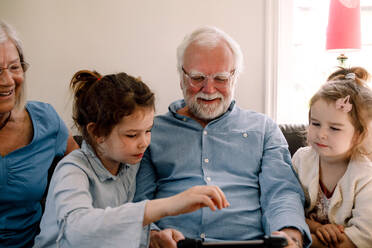 Smiling grandparents sitting with grandchildren while looking at digital tablet in living room at home - MASF13729