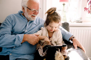 Girl using digital tablet while sitting with grandfather on sofa at home - MASF13733