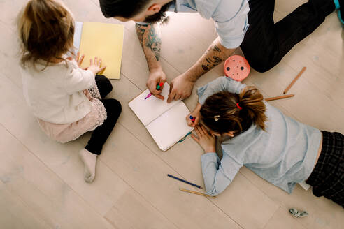 Hochformatige Ansicht eines Vaters, der seinen Töchtern beim Zeichnen auf dem Fußboden zu Hause hilft - MASF13740