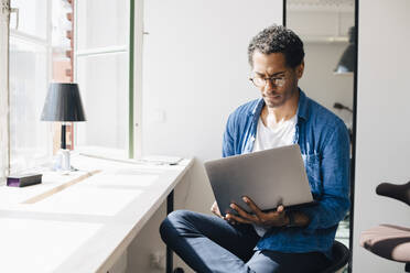 Computer programmer working on laptop while sitting by window in office - MASF13743