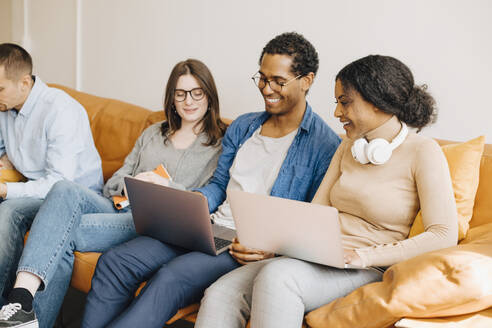 Lächelnde Programmierer, die über Laptops arbeiten, während sie auf dem Sofa in einem kreativen Büro sitzen - MASF13772