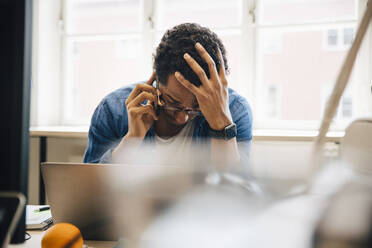 Frustrierter männlicher Computer-Hacker mit Hand in den Haaren, der mit einem Smartphone spricht, während er einen Laptop in einem kreativen Büro benutzt - MASF13787