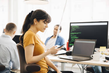 Female computer programmer using smart phone while sitting at desk in office - MASF13803