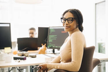 Porträt eines weiblichen Programmierers, der am Schreibtisch sitzt, mit einem Kollegen im Hintergrund in einem kreativen Büro - MASF13805