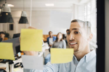 Male computer programmer sticking adhesive note on glass during meeting with colleagues in creative office - MASF13806