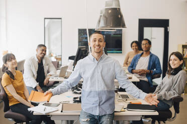 Portrait of smiling businessman gesturing while standing with colleagues sitting at desk in creative office - MASF13821