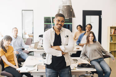 Portrait of confident computer hacker with arms crossed standing while happy colleagues sitting at desk in office - MASF13822