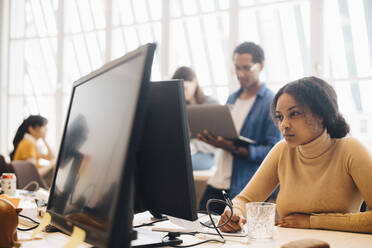 Konzentrierte Geschäftsfrau am Computer, während Kollegen im Hintergrund im Büro arbeiten - MASF13824