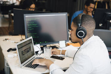 Seitenansicht eines männlichen Programmierers mit Kopfhörern, der über einem Laptop auf dem Schreibtisch programmiert, während er im Büro sitzt - MASF13836