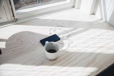 High angle view of smart phone and headphones with coffee cup on desk by window in office - MASF13841