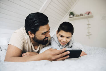 Father and smiling disabled son watching movie over mobile phone while lying on bed at home - MASF13886