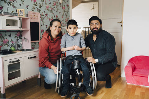 Porträt lächelnder Eltern, die zu Hause bei ihrem autistischen Sohn im Rollstuhl hocken, lizenzfreies Stockfoto