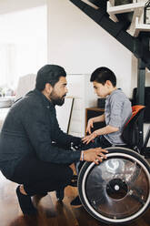 Side view of father looking at autistic son sitting on wheelchair at home - MASF13897