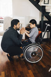 Side view of father holding autistic son's hands sitting on wheelchair at home - MASF13899