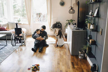 Hoher Blickwinkel des Vaters, der seinen autistischen Sohn hält, während seine Tochter zu Hause auf dem Boden sitzt - MASF13901