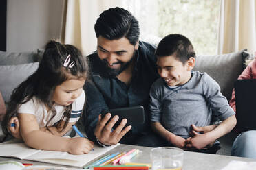 Father showing mobile phone to children while woman using laptop on sofa in living room - MASF13905